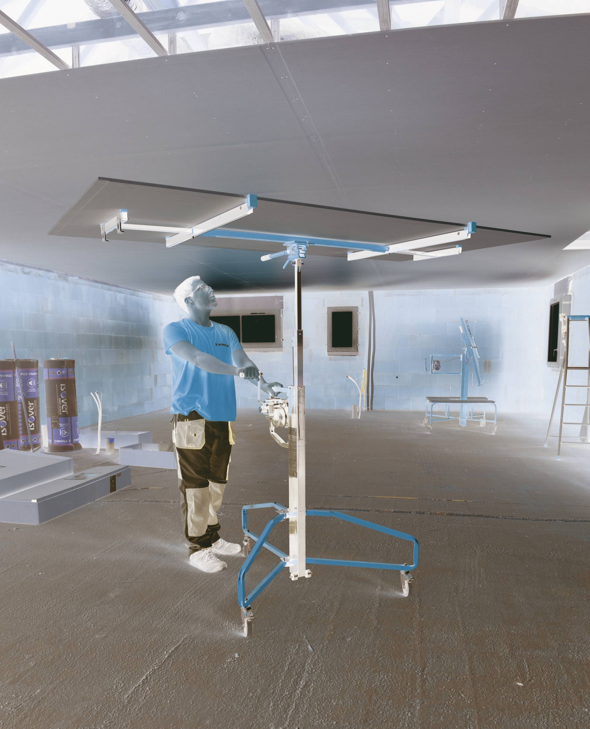 Un ouvrier pulvérise ou peint un grand plafond intérieur à l'aide d'un échafaudage mobile dans une pièce largement vide en cours de construction ou de rénovation.