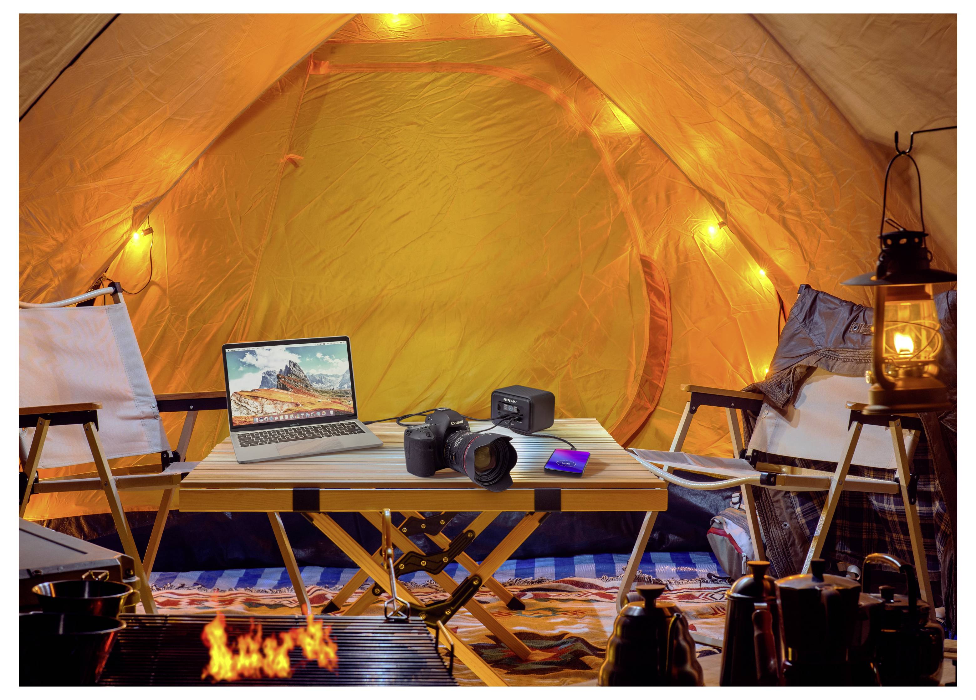 Intérieur d'une tente de camping chaleureusement éclairé avec un ordinateur portable, un appareil photo, un sac à dos, des chaises, des lanternes et un petit feu devant un espace de table aménagé.