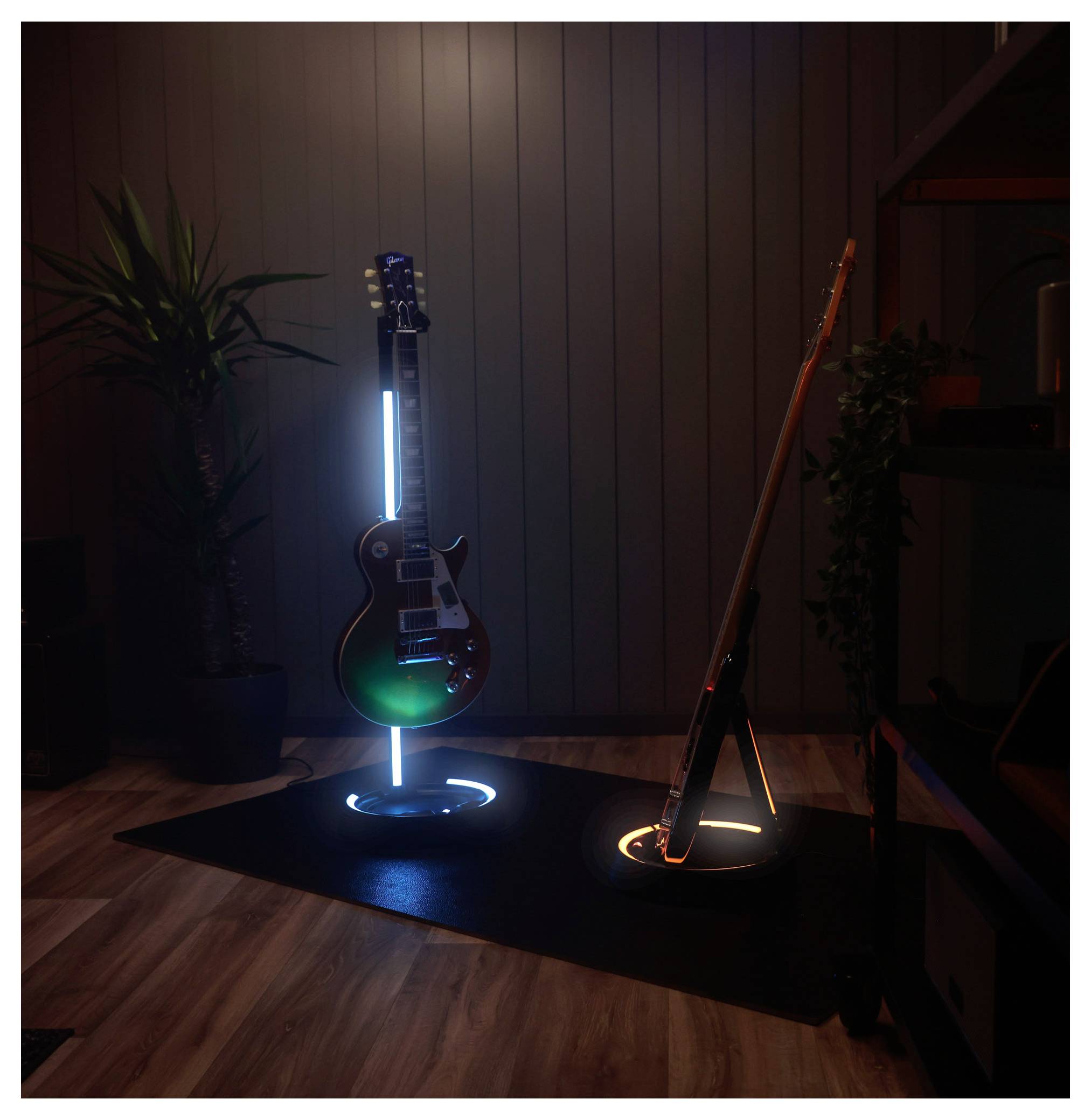Deux guitares électriques sur des supports de sol illuminés dans une pièce sombre, l'une éclairée en bleu et l'autre en orange, avec des plantes et du lambris en bois en arrière-plan.