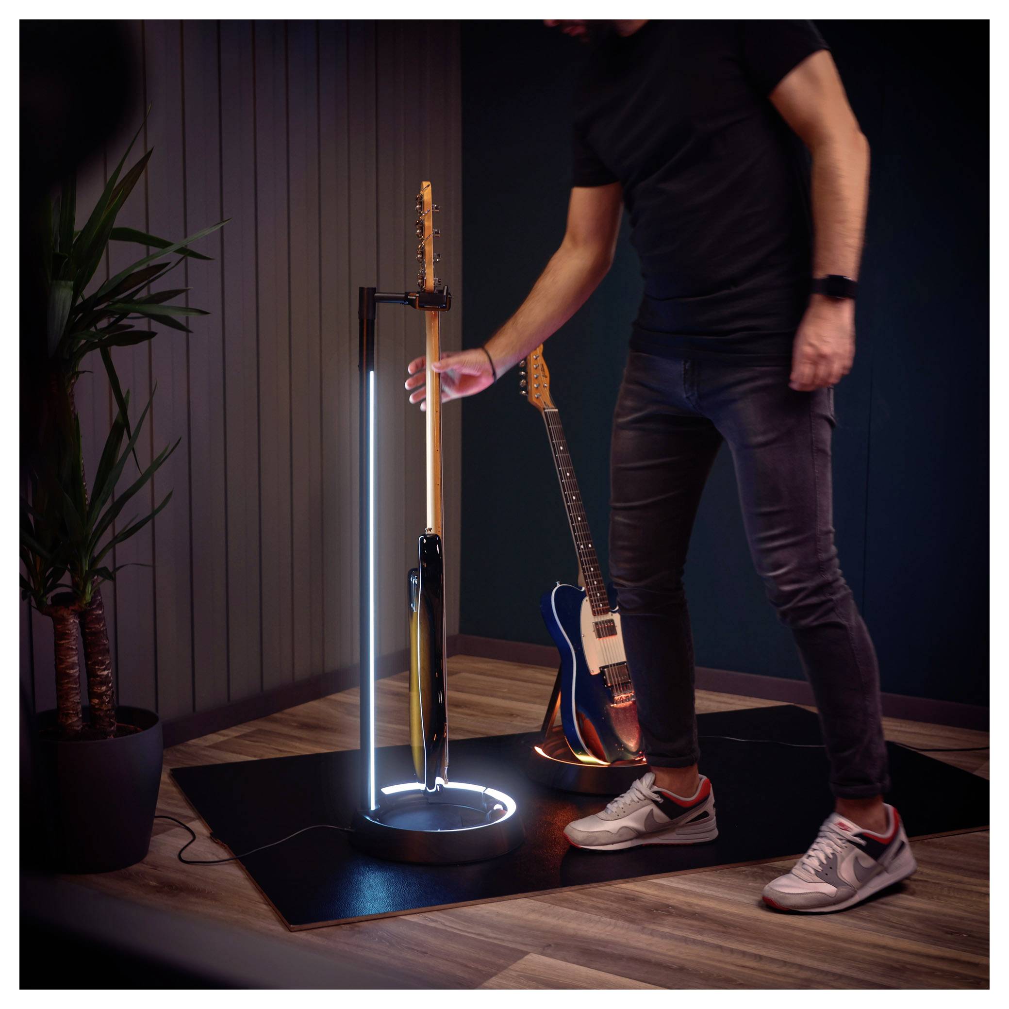 Personne ajustant un support de sol luminescent tenant des guitares dans une pièce moderne avec un sol en bois et une plante en pot.