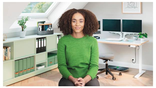 Une femme dans un pull vert est assise en souriant dans un bureau à domicile avec des moniteurs doubles, des étagères et un bureau derrière elle.