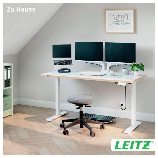 Bureau à domicile avec bureau réglable, deux moniteurs, ordinateur portable, tabouret, clavier et plante sous un cadre mural; 'Zu Hause' et logo LEITZ visibles.