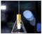 Luminaire suspendu moderne avec un abat-jour en verre transparent de forme conique et une partie supérieure dorée, suspendu devant un arrière-plan flou et sombre.