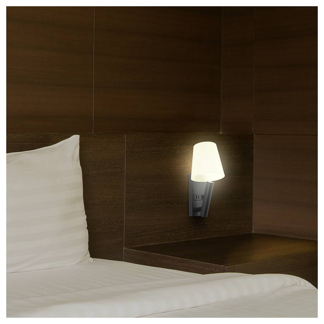 Gros plan d'un lit soigneusement fait avec un oreiller blanc et une applique murale illuminée dans une chambre à panneaux de bois foncé.