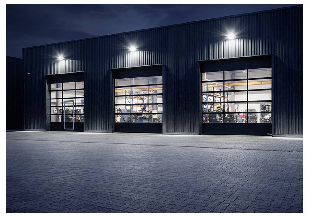 Extérieur nocturne d'un entrepôt industriel avec trois portes de garage en verre illuminées, montrant l'équipement à l'intérieur et une cour pavée vide.