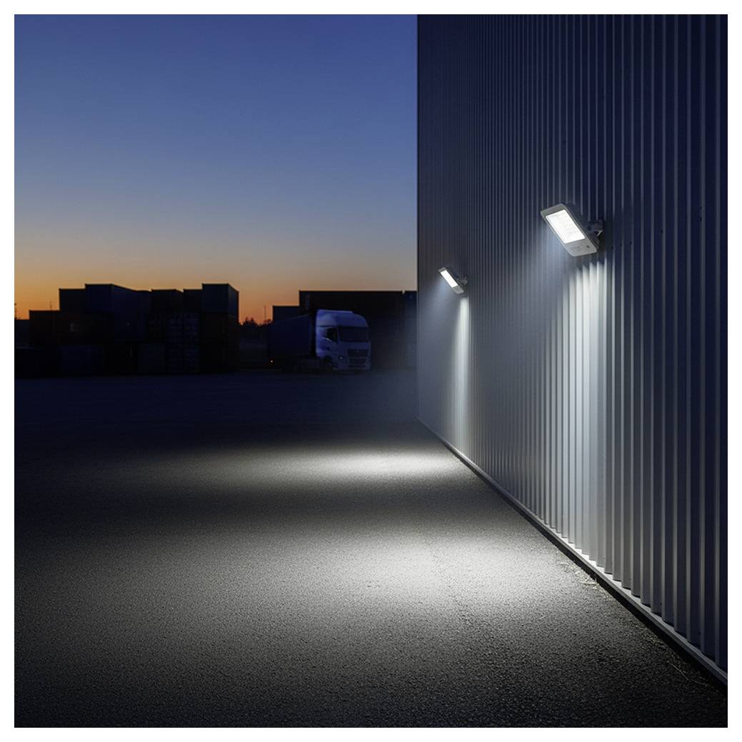 Extérieur d'un entrepôt au crépuscule, avec des luminaires muraux illuminant le revêtement en métal ondulé et le parking pavé vide.