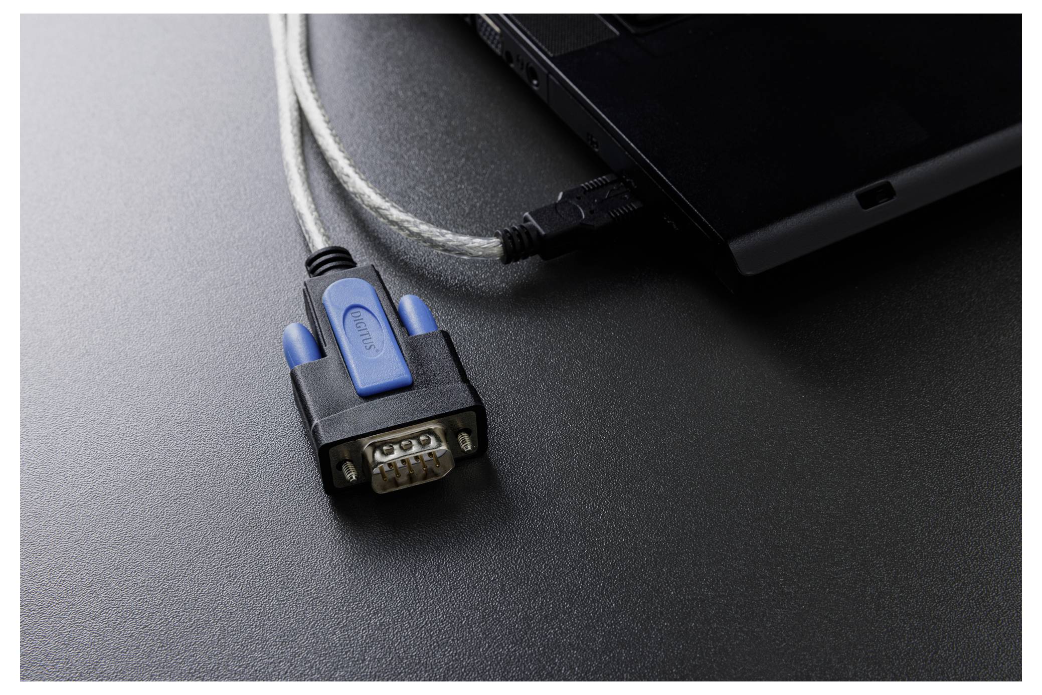 Gros plan d'un ordinateur portable avec un adaptateur série vers USB et un câble branchés sur une surface sombre.