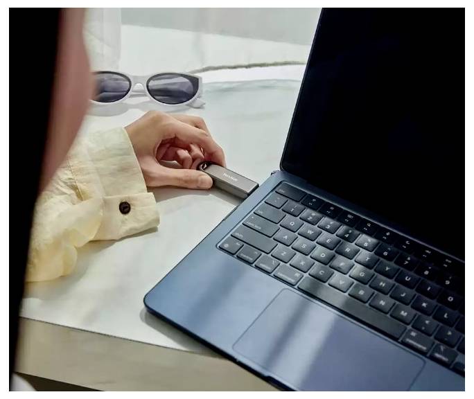 Une personne branche une clé USB dans un ordinateur portable à côté de lunettes de soleil sur un bureau.