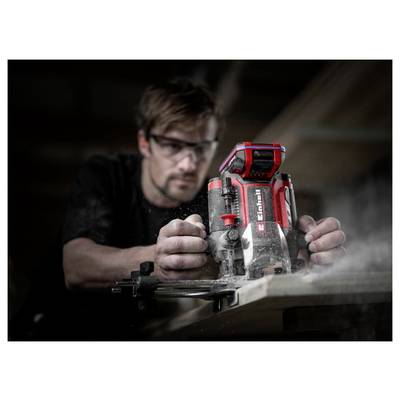 Un homme portant des lunettes de sécurité utilise un outil électrique portatif rouge pour couper du bois, envoyant de la sciure dans l'air dans un atelier.