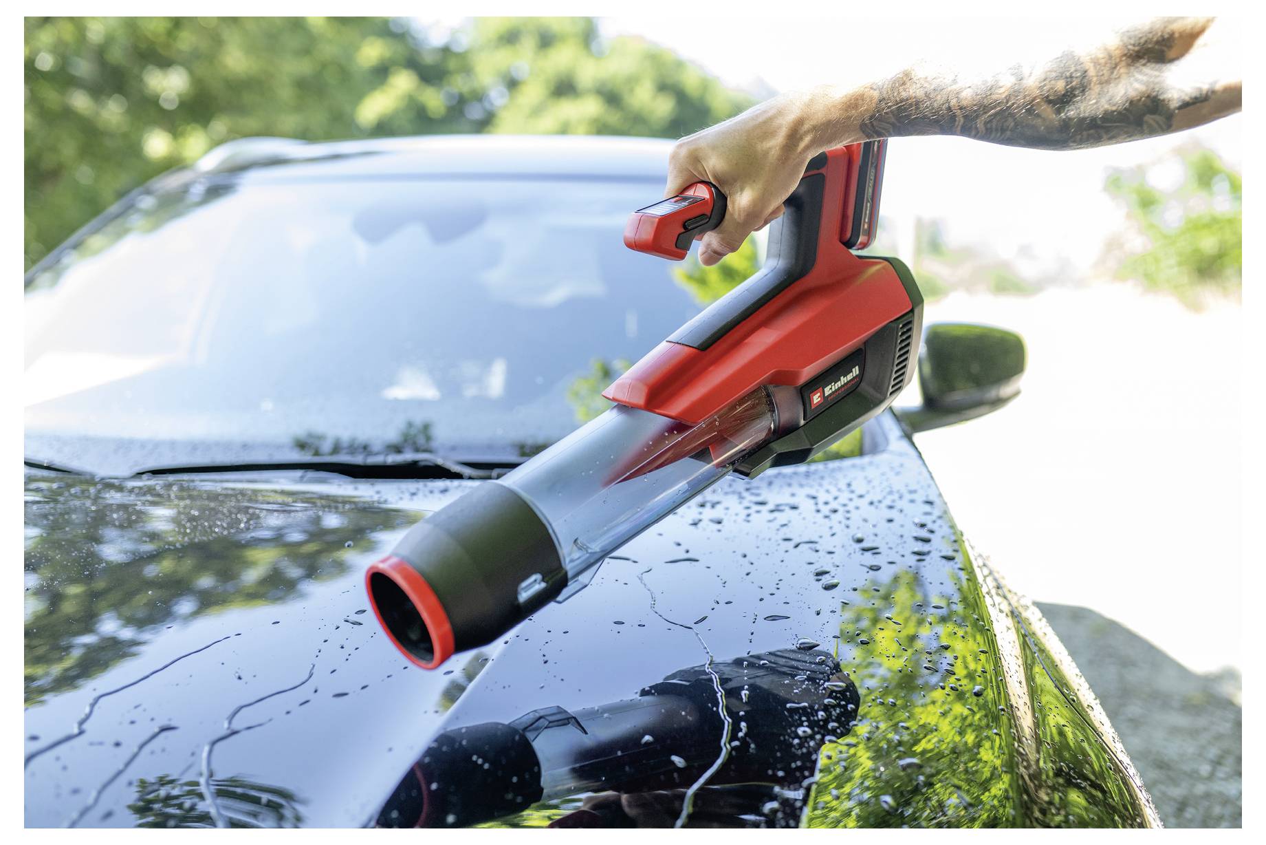 Une personne utilisant un souffleur à feuilles sans fil rouge pour dégager l'eau et les débris du capot mouillé d'une voiture à l'extérieur.