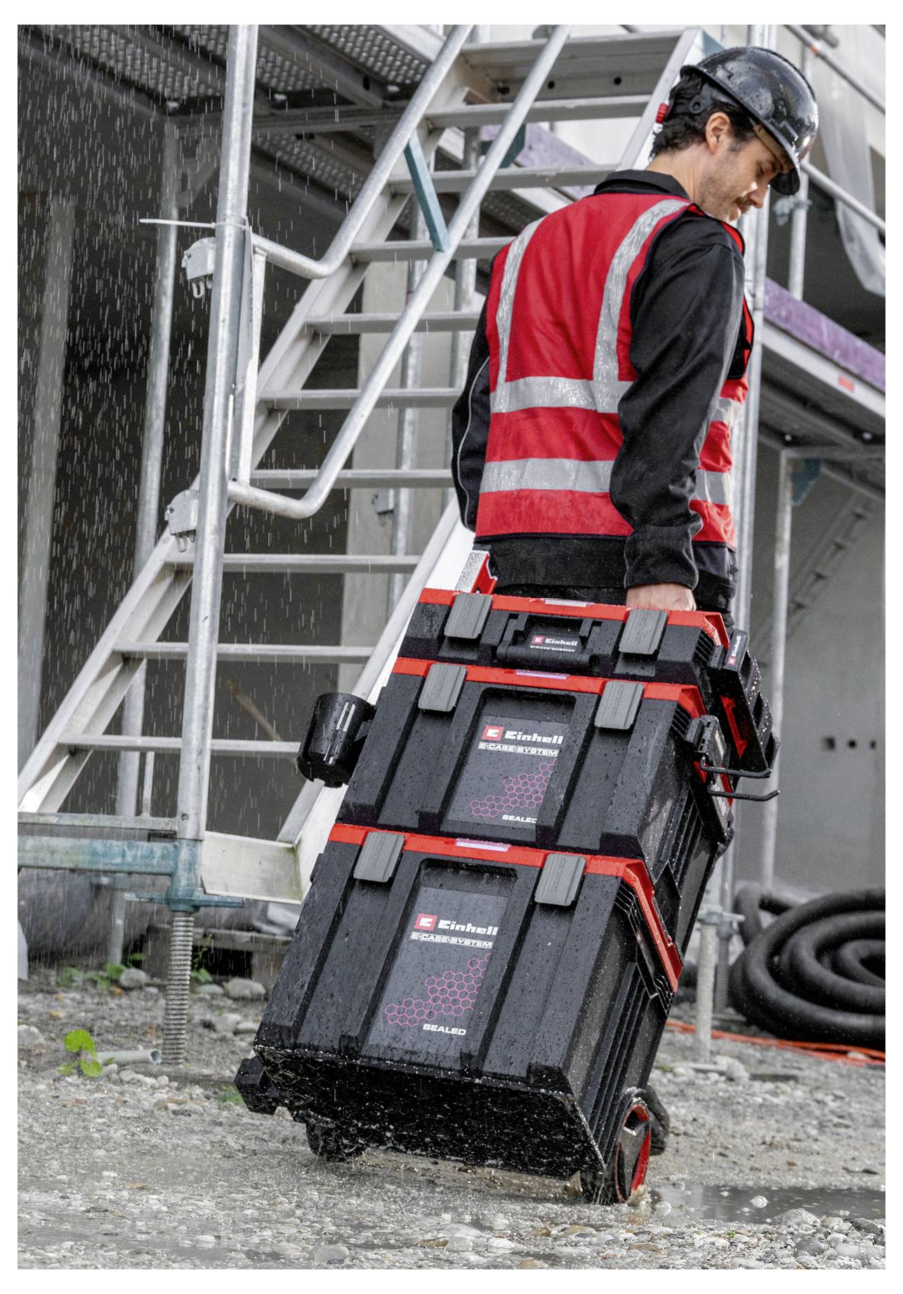 Un ouvrier du bâtiment portant un casque de protection et un gilet haute visibilité transporte deux caisses à outils empilées sur un chantier industriel à côté d'escaliers en métal.