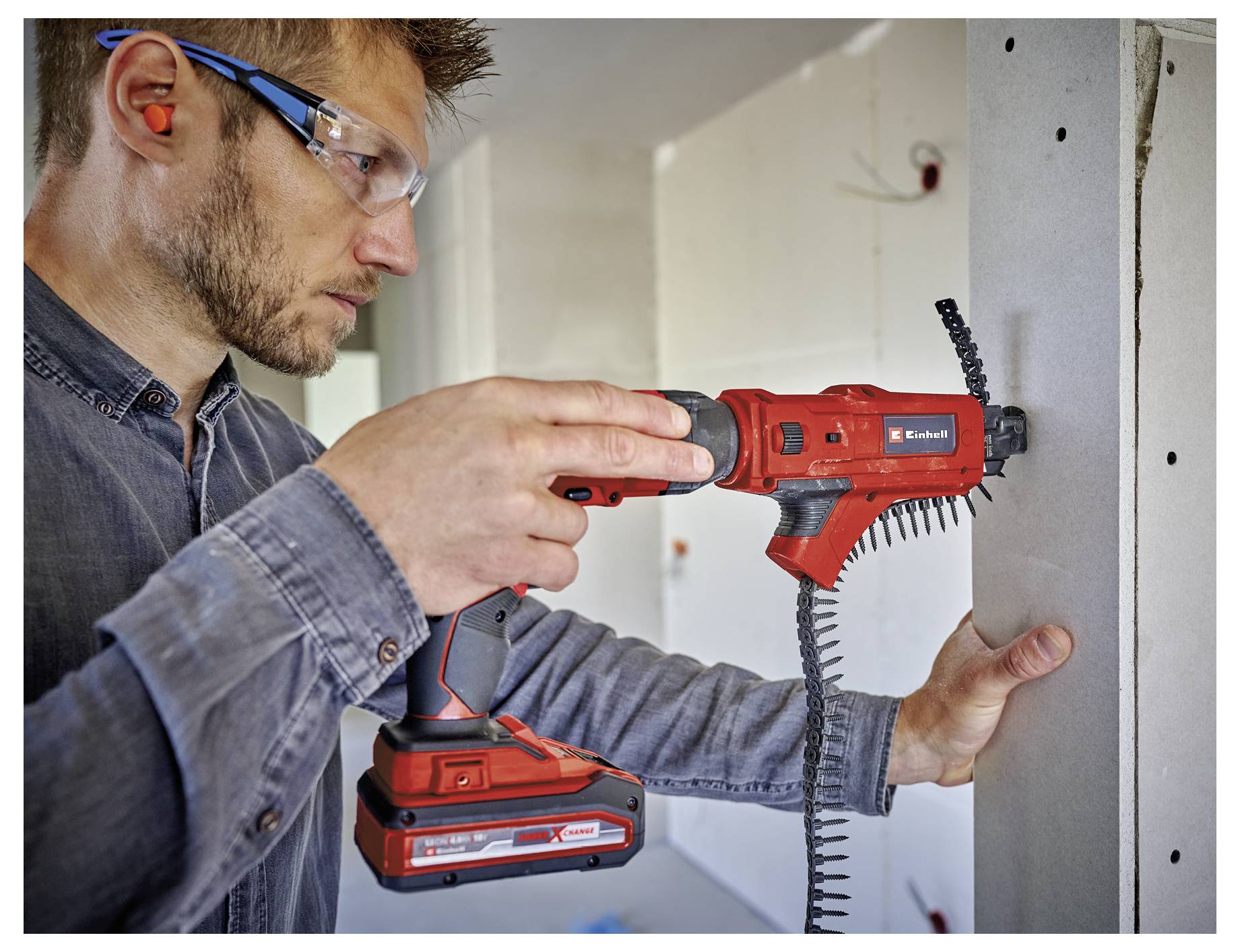 Ouvrier du bâtiment utilisant une perceuse-visseuse sans fil rouge pour fixer un panneau mural tout en portant des lunettes de sécurité et des bouchons auriculaires.