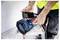 Une personne vêtue d'un gilet haute visibilité utilise un smartphone pour charger ou connecter un haut-parleur portable bleu sur un établi.