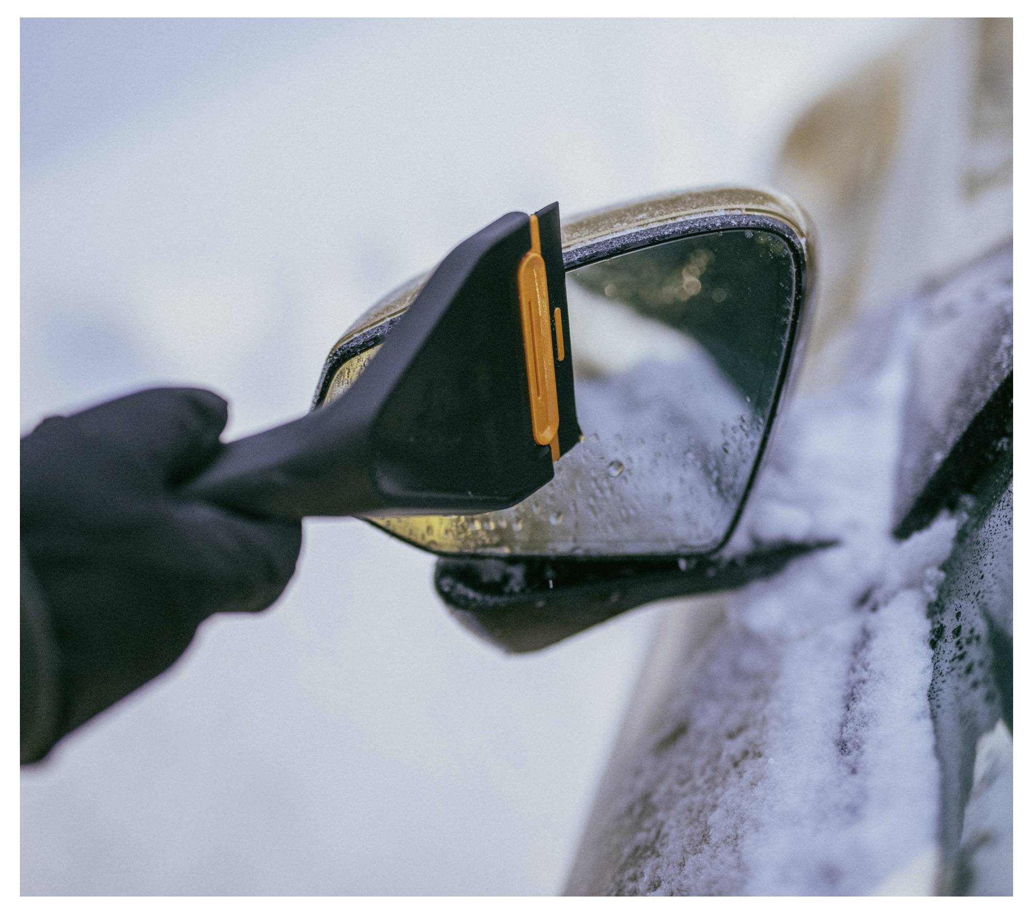 Une main gantée gratte la glace d'un rétroviseur latéral de voiture avec un grattoir à glace sur un véhicule enneigé.