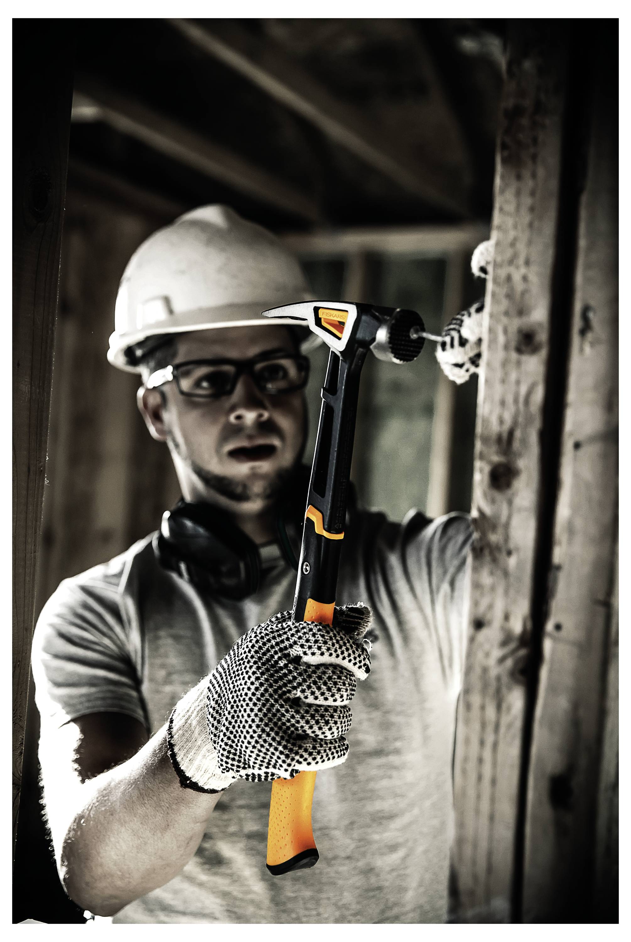 Ouvrier du bâtiment portant un casque de protection et des gants tenant un marteau, travaillant à l'intérieur d'une charpente de bâtiment.