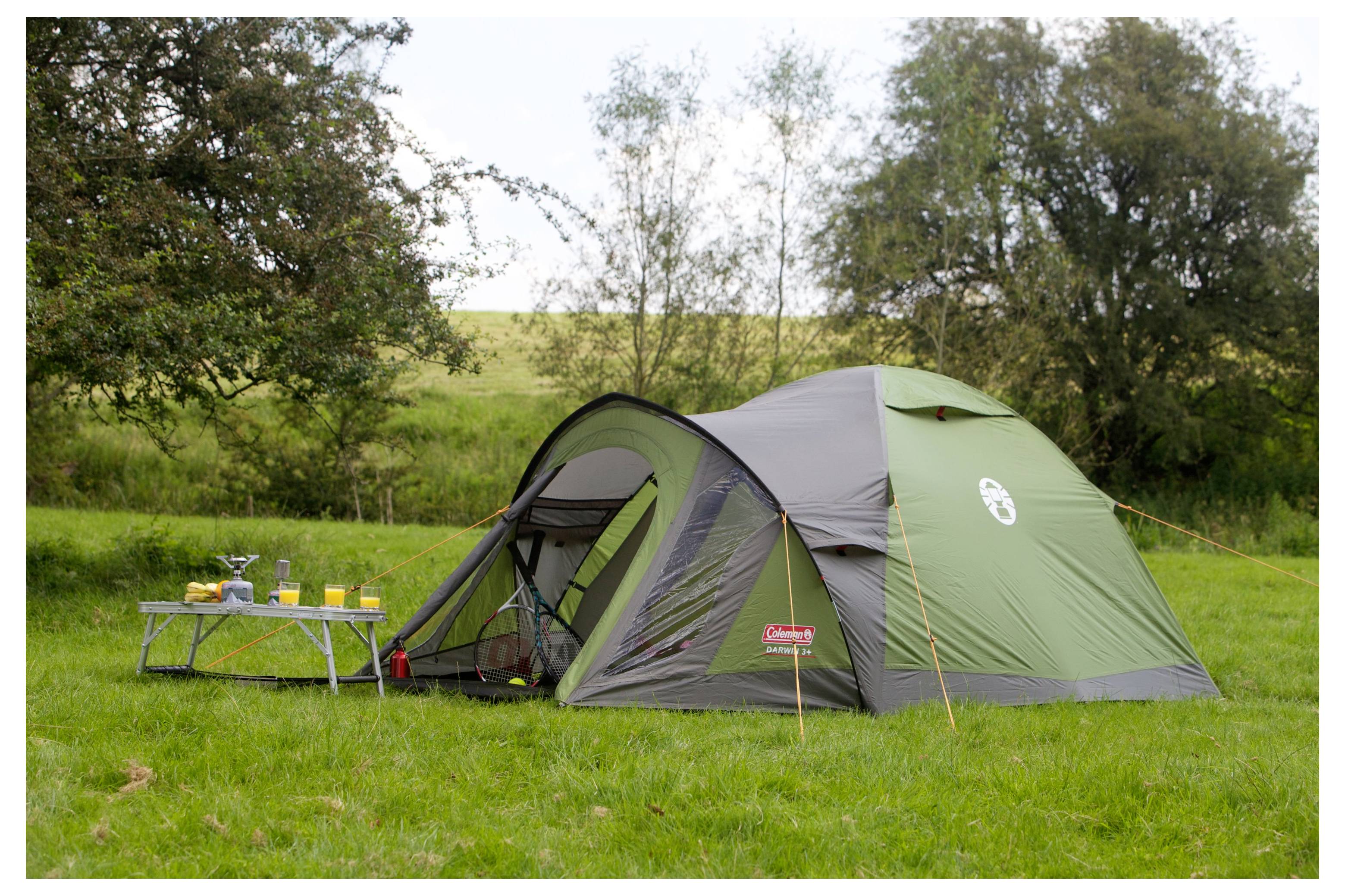 Tente de camping verte dressée sur un champ herbeux à côté d'une petite table avec des boissons et du matériel de camping, entourée d'arbres et de la campagne ouverte.