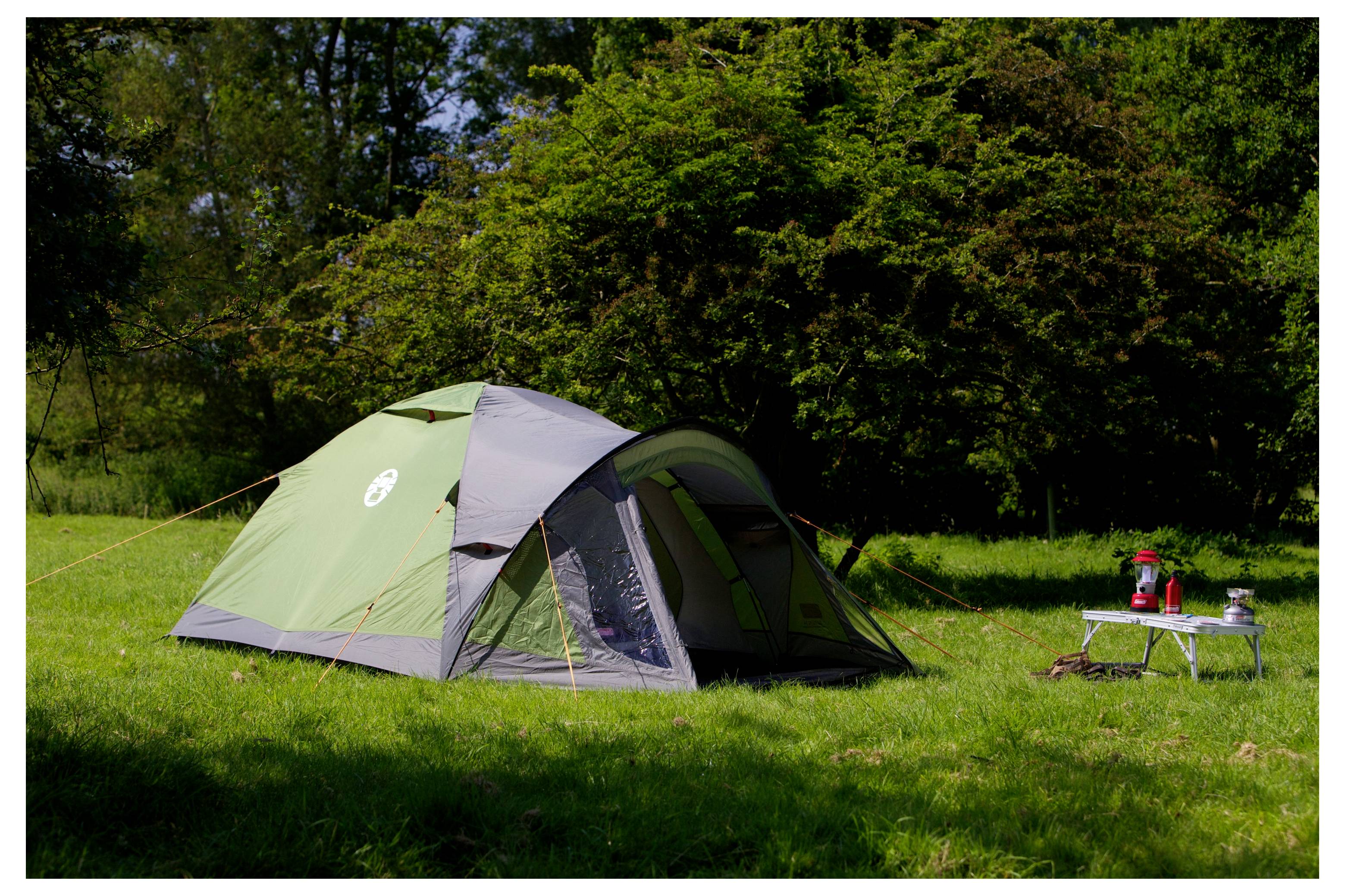 Tente dôme verte dressée sur un terrain de camping herbeux à côté d'une petite table avec du matériel de camping, avec des arbres en arrière-plan.