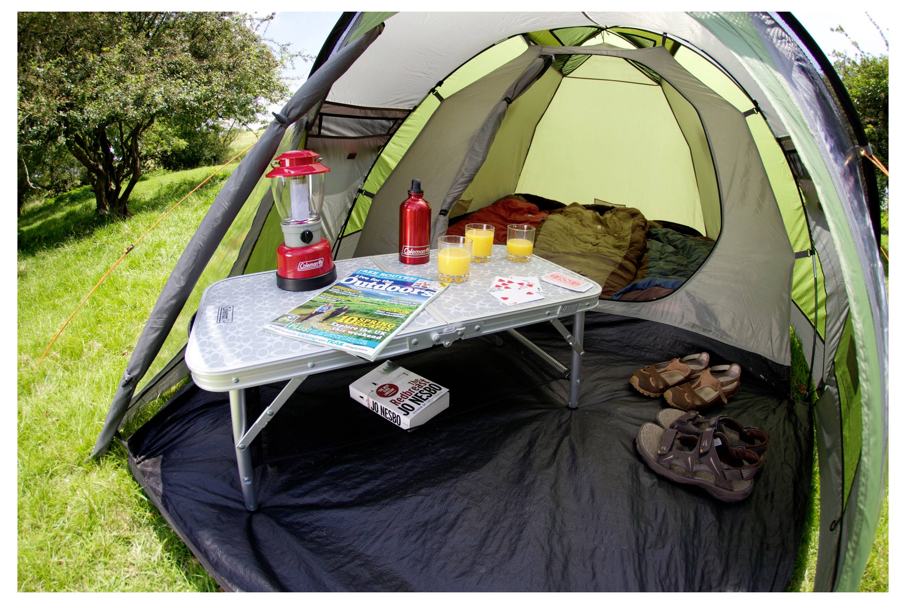 Une tente de camping installée sur l'herbe avec une table pliante, une lanterne, des boissons, un magazine, un jeu de cartes, des chaussures et des sandales à l'intérieur.