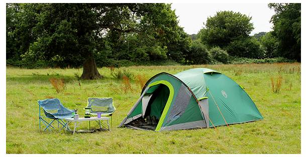 Configuration de camping dans un champ herbeux avec une tente dôme verte, deux chaises pliantes et une petite table à côté des arbres.