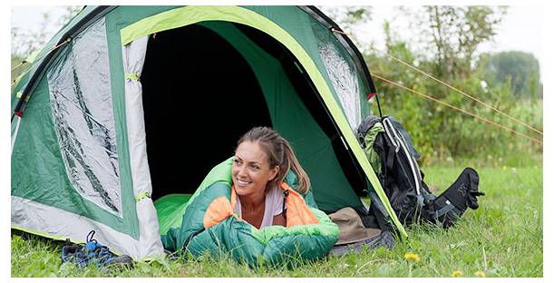 Femme souriante allongée dans un sac de couchage vert à l'entrée d'une tente de camping sur l'herbe, avec son sac à dos et son équipement à côté d'elle.