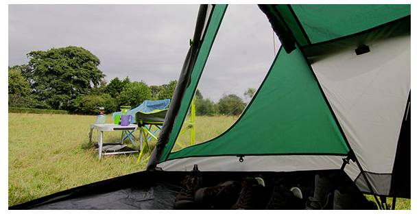 Vue de l'intérieur d'une tente de camping verte donnant sur un champ herbeux avec une table pliante, des chaises et des arbres sous un ciel couvert.