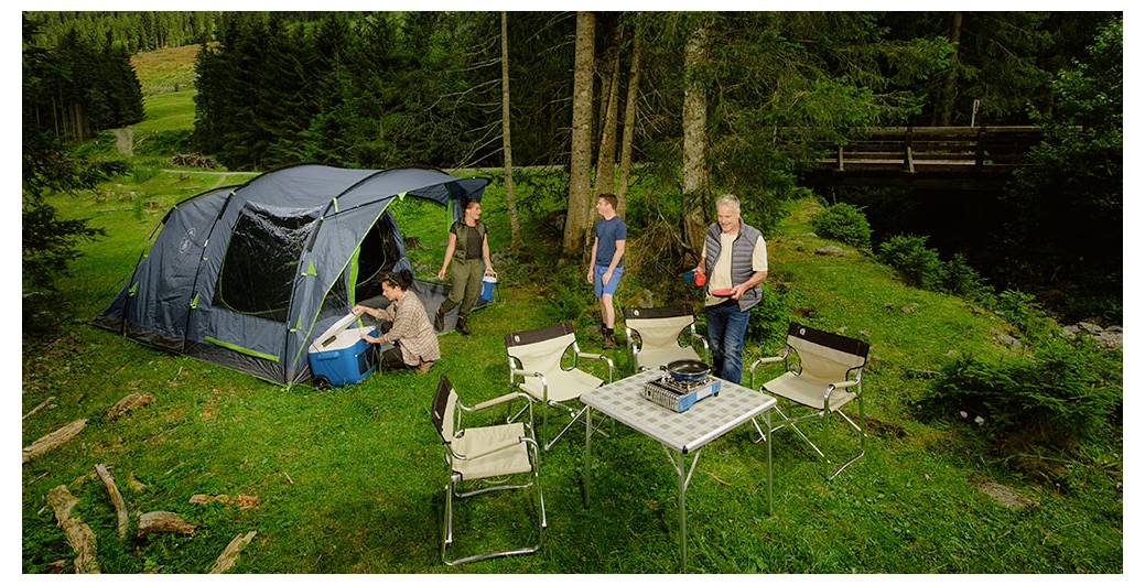 Des personnes campant à côté d'une grande tente sur une rive herbeuse, avec des chaises pliantes et une table au premier plan.