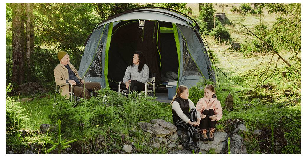 Une famille se détend sur un terrain de camping en dehors d'une tente dôme verte, avec deux personnes assises à l'intérieur et deux autres assises sur des rochers devant, entourées d'arbres et de feuillage.