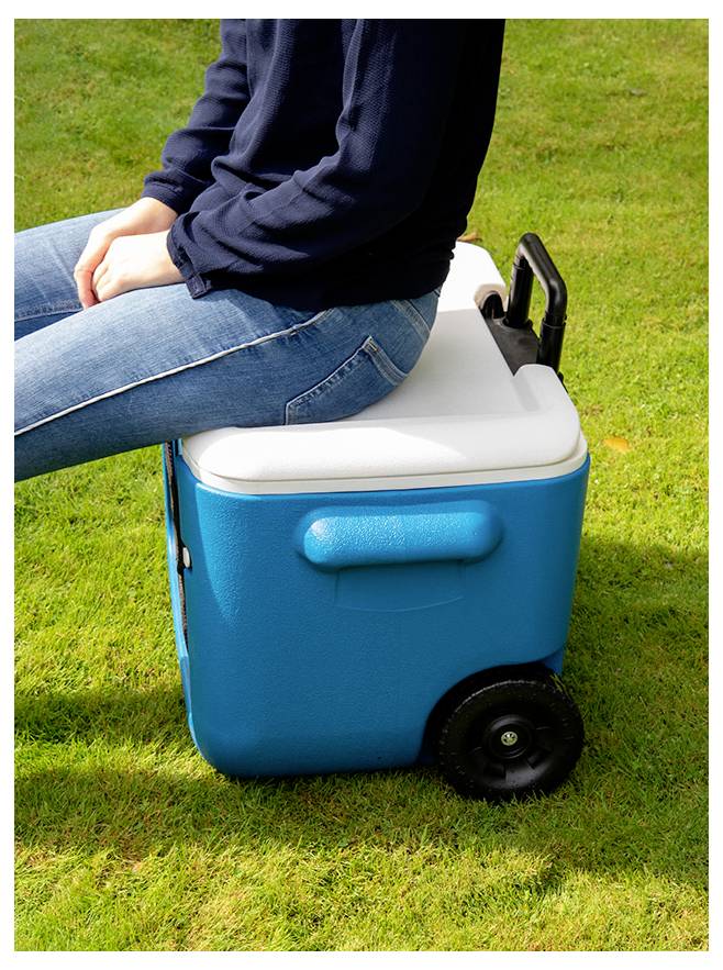 Personne assise sur une glacière bleue avec roues sur l'herbe à l'extérieur, l'utilisant comme siège et chariot portable.