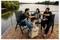 Trois amis se détendent sur une terrasse en bord de rivière, souriants et trinquant avec des verres tout en étant assis dans des chaises à côté d'une glacière.