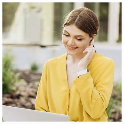 Femme souriante en blouse jaune ajuste son écouteur tout en regardant un ordinateur portable à l'extérieur.