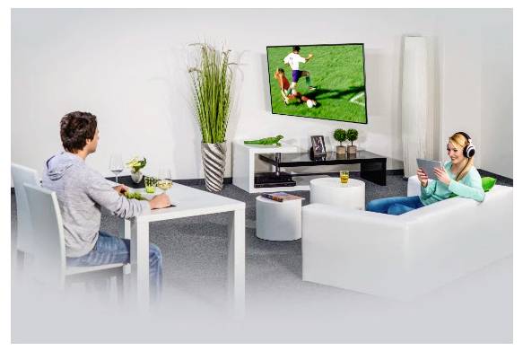 Deux personnes se détendent dans un salon en regardant un match de football sur un téléviseur fixé au mur, l'une assise à une table et l'autre sur un canapé utilisant une tablette.