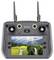 Télécommande de drone grise avec deux antennes, manettes de jeu, bouton d'alimentation et écran intégré affichant un flux vidéo en direct du paysage montrant des montagnes, des champs et une rivière.