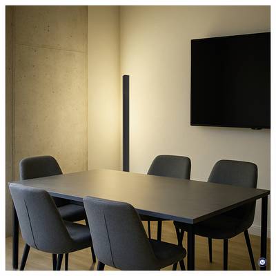 Salle de réunion moderne avec une table rectangulaire, quatre chaises, un téléviseur mural et un lampadaire contre des murs en béton et blancs.