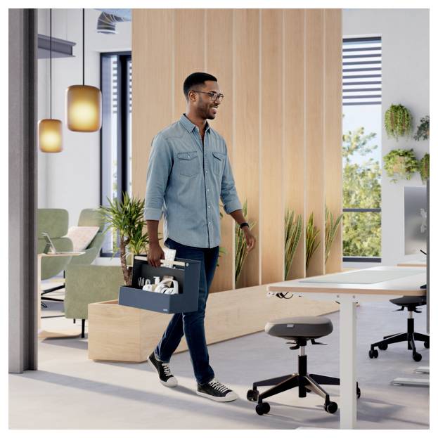 Un homme portant une boîte à outils traverse un espace de travail de bureau moderne avec des bureaux, des chaises et des plantes.