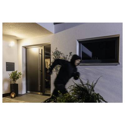 Intrus masqué vêtu de noir qui court devant l'entrée illuminée d'une maison la nuit, avec la porte d'entrée ouverte et la fenêtre sombre.