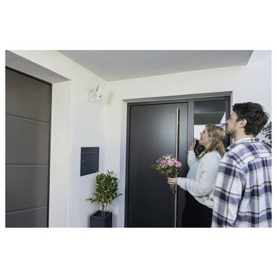 Un couple se tient devant la porte d'un appartement moderne, la femme tenant des fleurs roses et sonnant à la porte.
