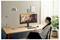 Une femme est assise à un bureau en tenant une tasse, regardant un moniteur affichant une fille avec un chien ; un bureau à domicile avec une lampe, un clavier et une plante.
