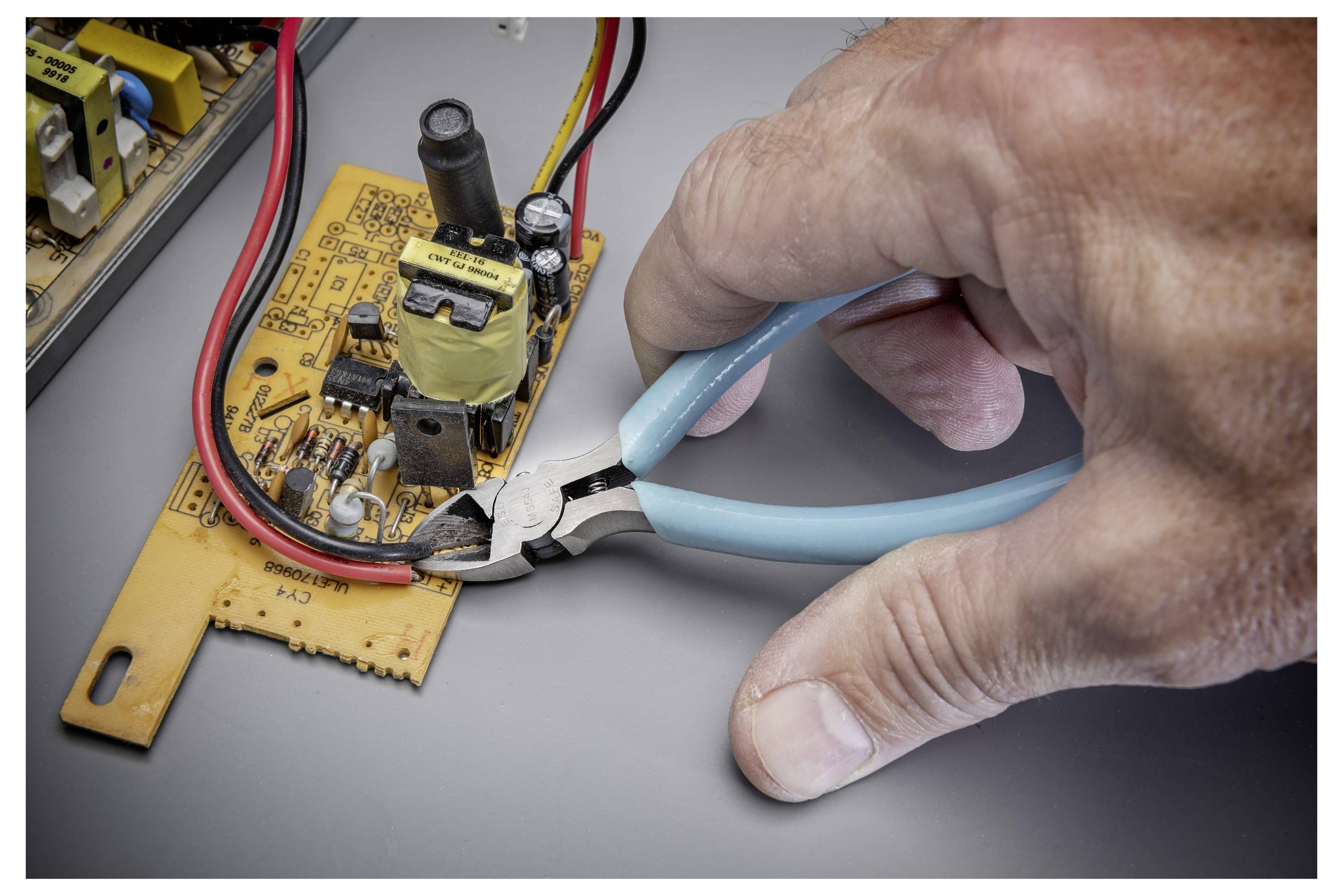 Une main utilisant des coupe-fils bleus pour couper un fil sur une petite carte de circuit imprimé jaune lors de la réparation électronique.