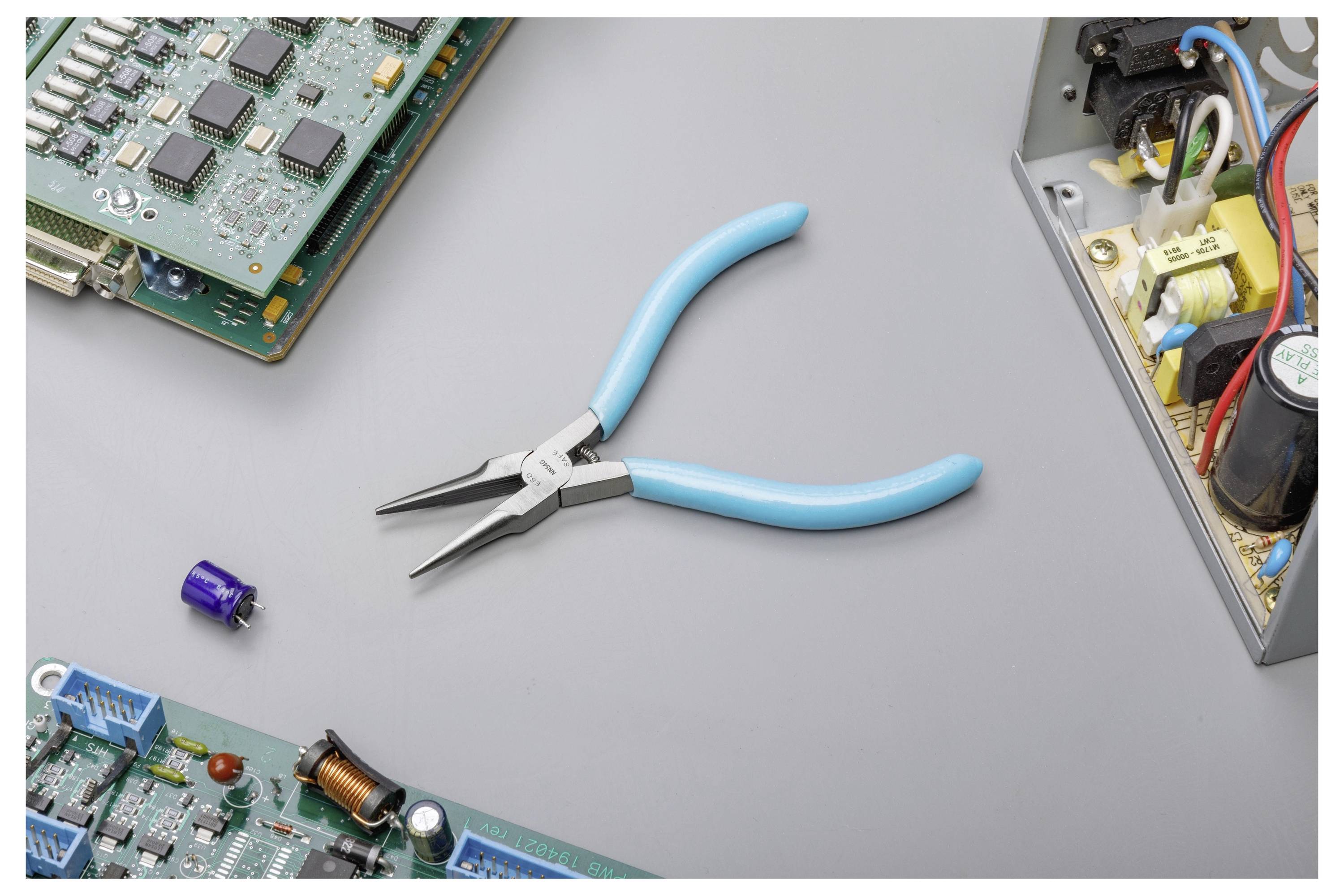 Pince à bec effilé à poignée bleue sur une surface de travail grise, entourée de cartes de circuits électroniques désassemblées et de composants.
