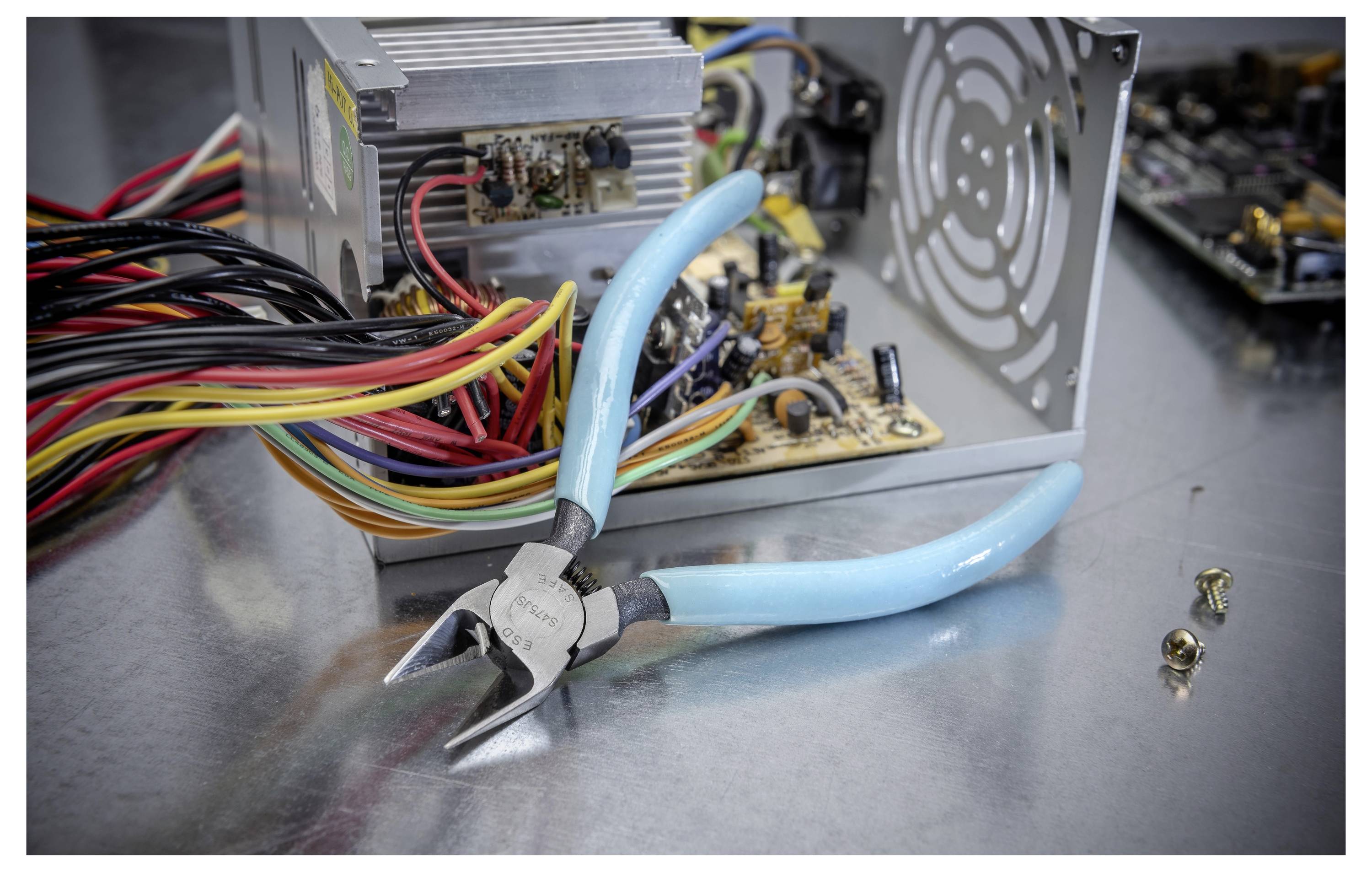 Ciseaux à fils à poignée bleue devant une alimentation électrique d'ordinateur ouverte avec carte de circuit exposée et fils groupés sur une surface métallique.