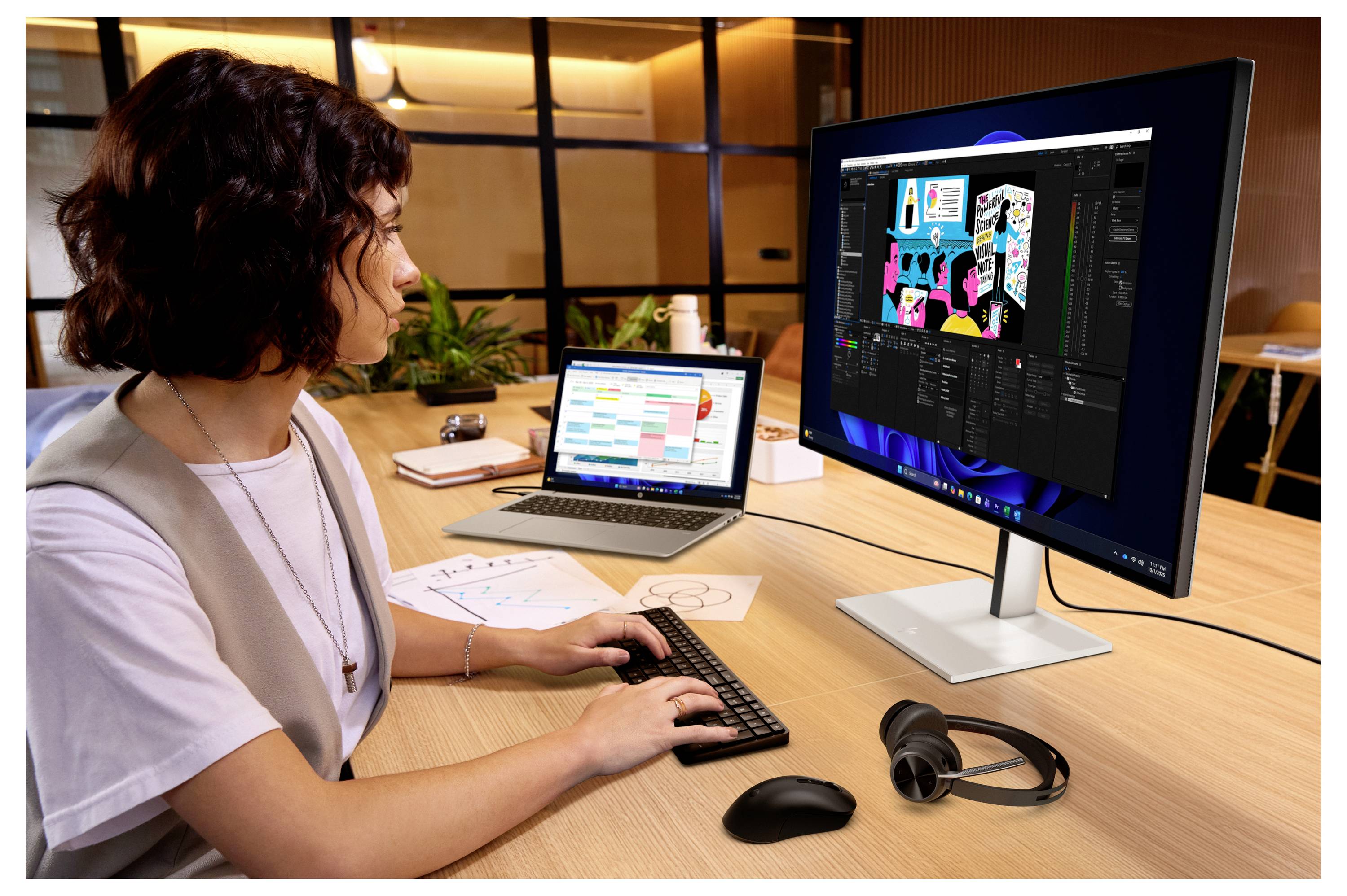 Femme à un bureau qui conçoit des graphiques sur un ordinateur portable et un grand moniteur, en utilisant une souris et un clavier dans un espace de travail de bureau moderne.