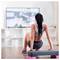 Femme qui fait de l'exercice à la maison en regardant une vidéo d'entraînement à la télévision, assise sur un banc de step dans un salon lumineux.