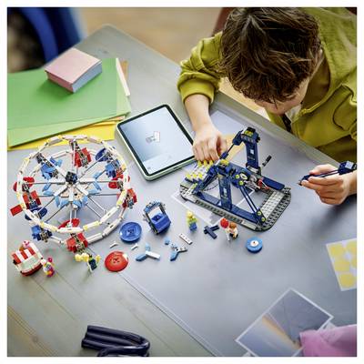 Un enfant construisant une grande roue LEGO et une grue à une table, avec des pièces dispersées, une tablette et d'autres matériaux de bricolage autour.