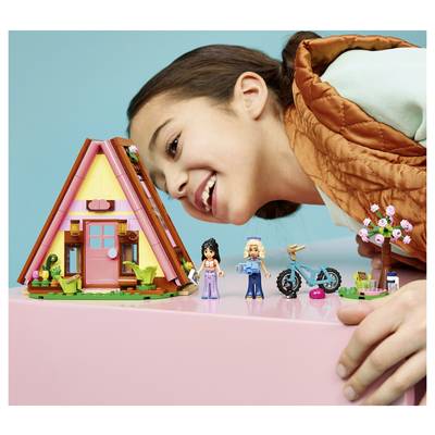 Un enfant souriant se penche vers une maison-A miniature en bois avec des poupées minuscules, un vélo et des fleurs sur une table rose sur un fond bleu.