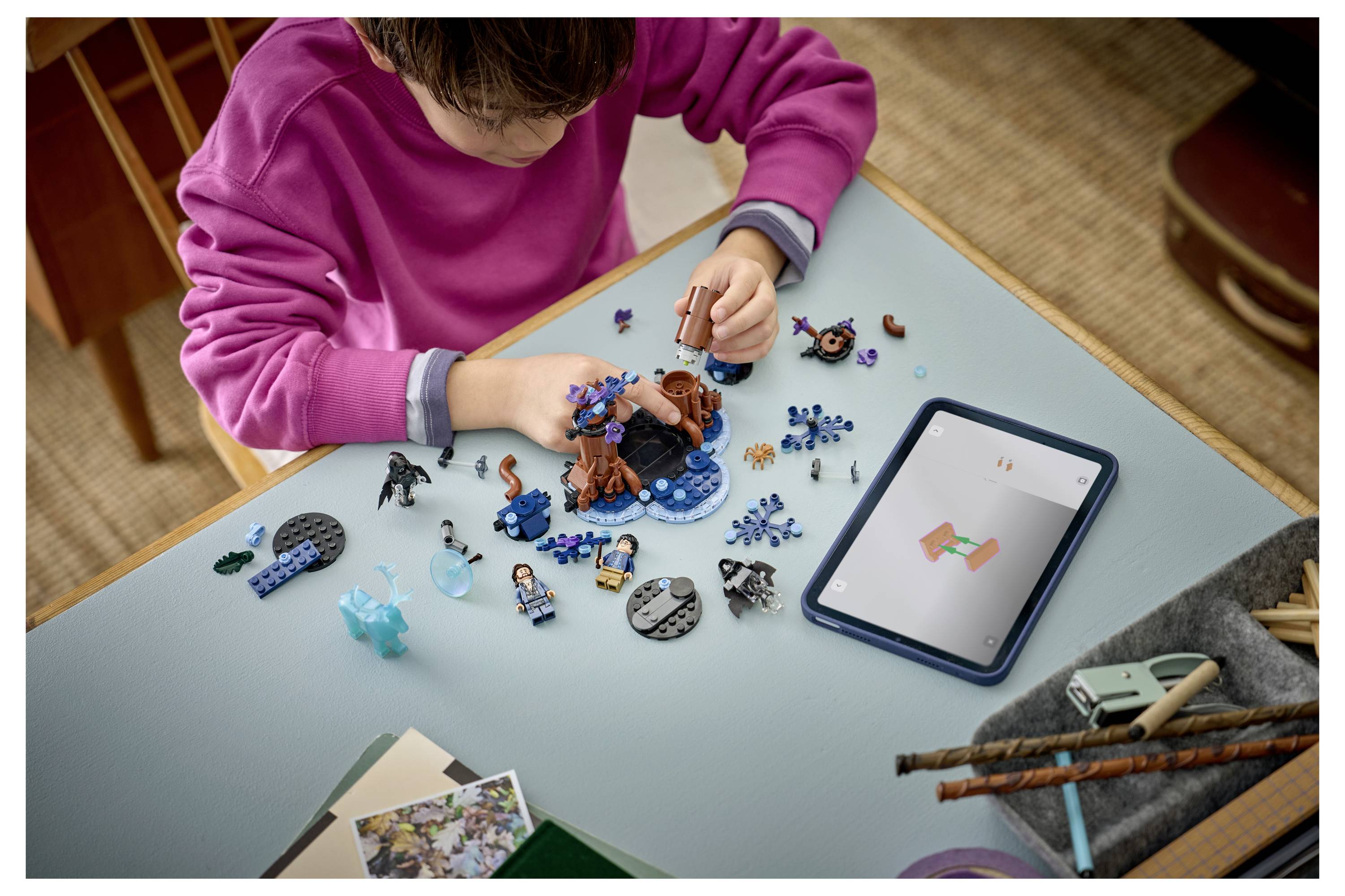 Un enfant dans un pull rose construisant une scène de jouets avec de petites figurines et des pièces sur une table à côté d'une tablette affichant un modèle 3D.