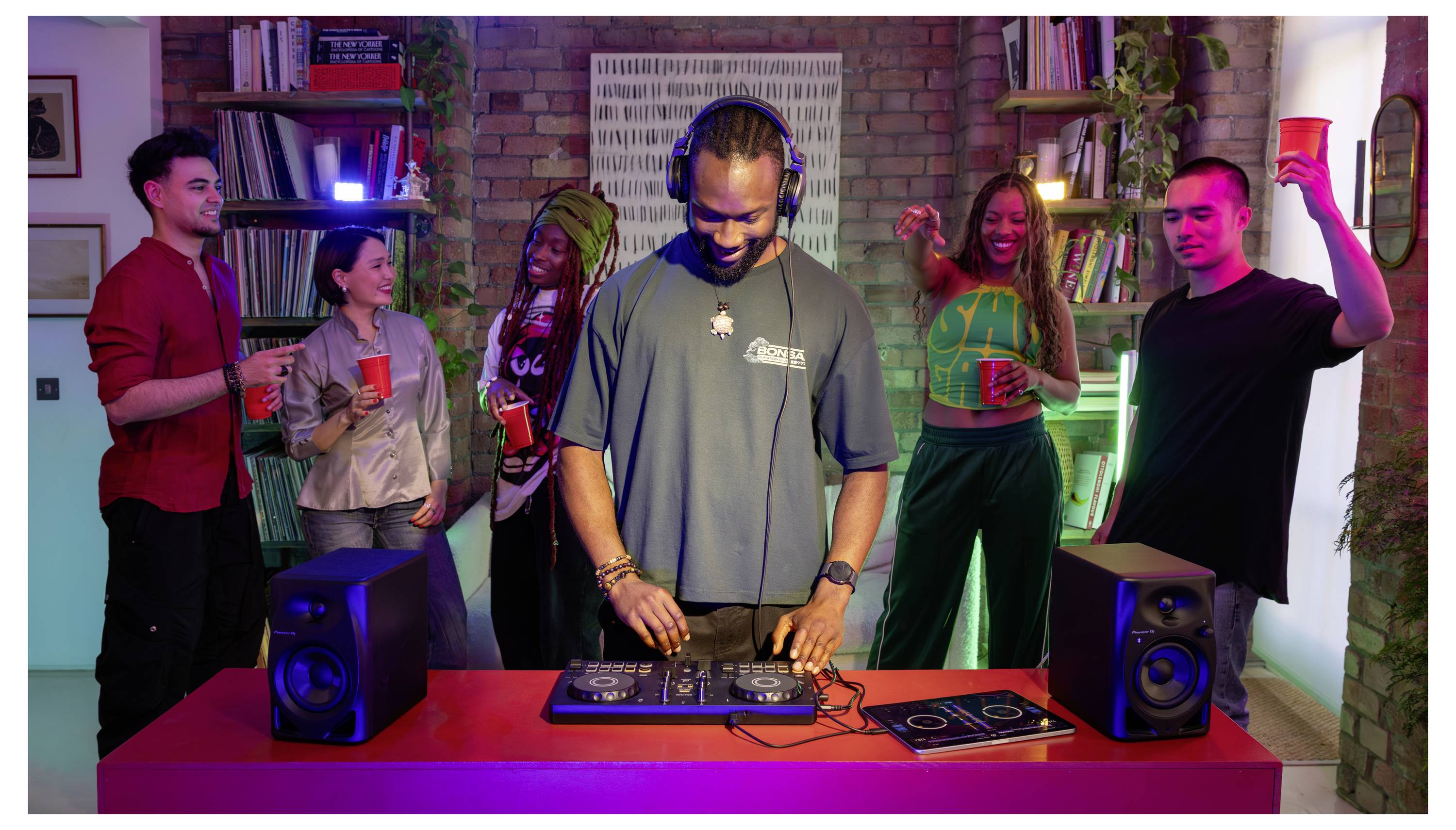 Un DJ portant des écouteurs mixe de la musique à une table tandis que cinq personnes se tiennent autour de lui en tenant des verres dans une scène de fête colorée en intérieur.