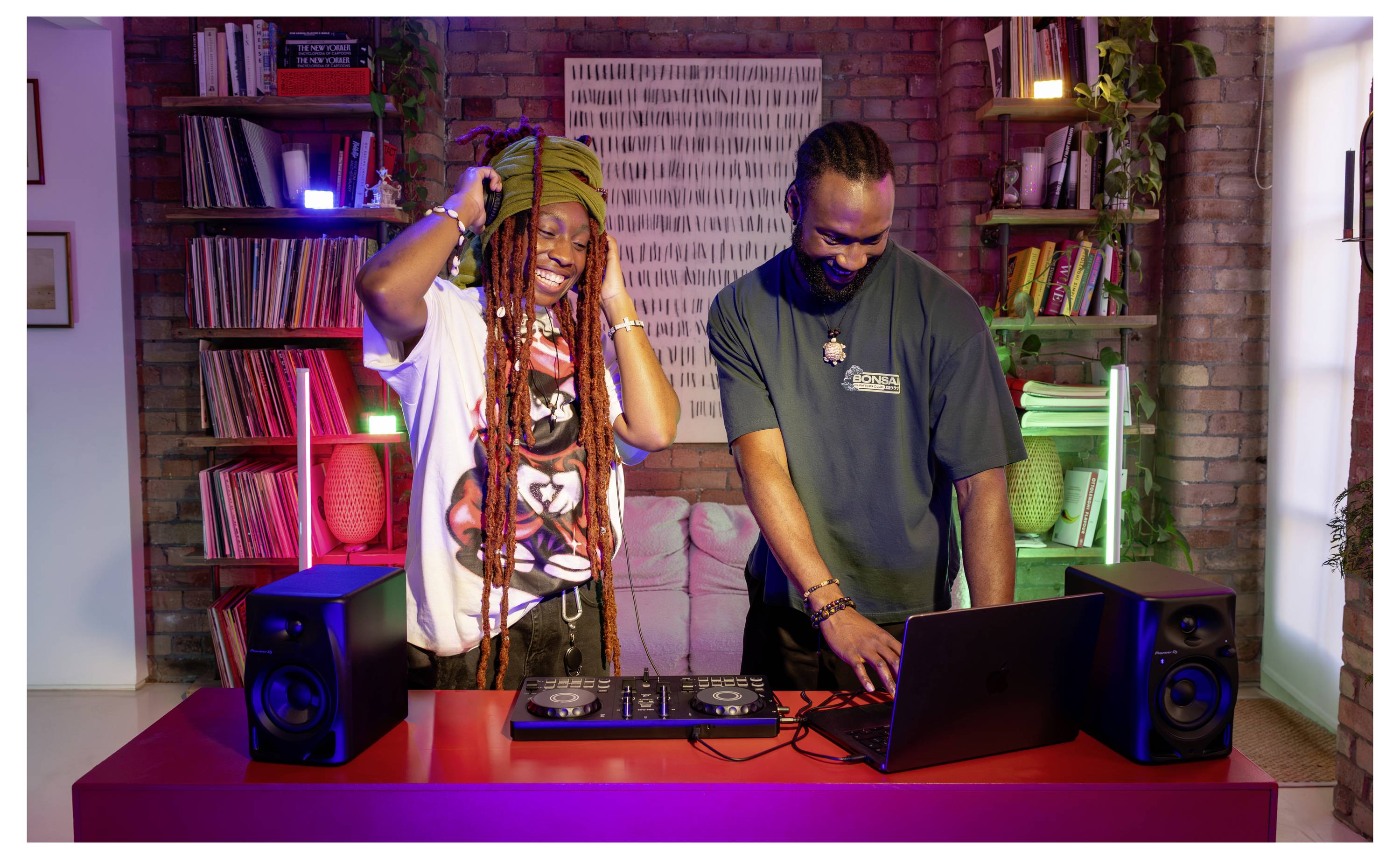 Deux DJ se tiennent devant une platine vinyle et un ordinateur portable dans un studio coloré, l'un sourit avec un casque audio tandis que l'autre travaille sur la musique.
