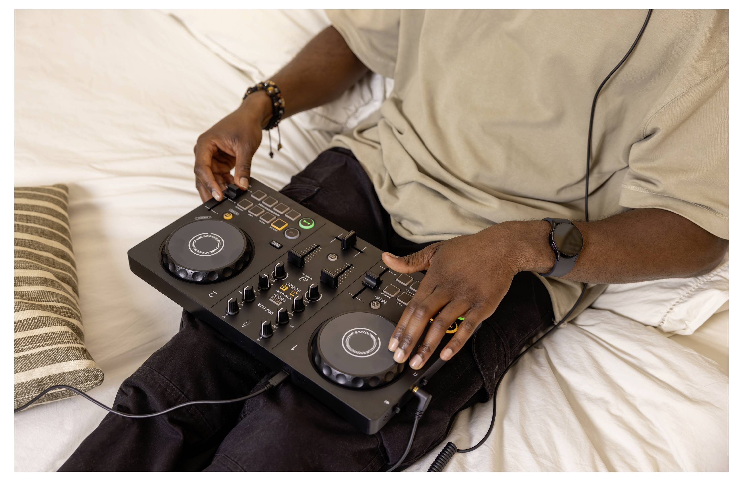 Personne utilisant un contrôleur DJ compact sur ses genoux en étant assise sur un canapé, les mains sur les molettes de navigation et les commandes du mélangeur.