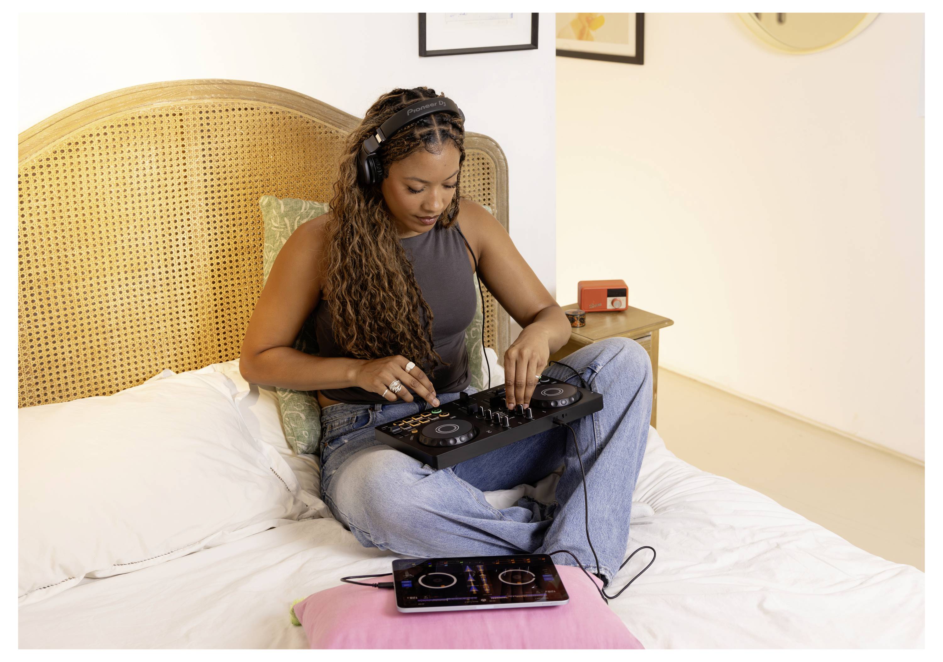 Femme assise sur un lit utilisant une contrôleuse DJ, avec une tablette ou un téléphone à côté d'elle connecté par un câble.