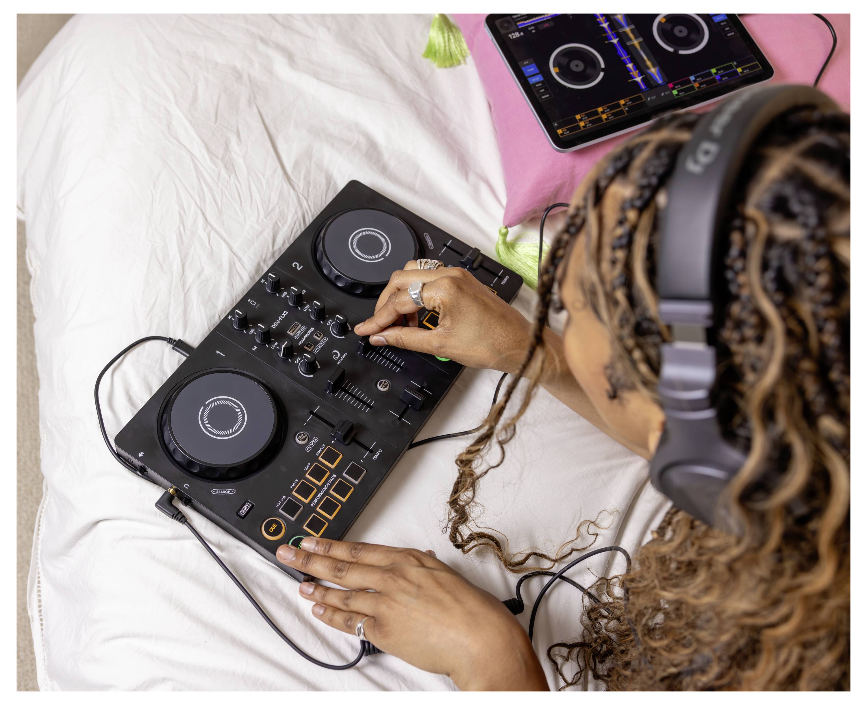 Personne portant des écouteurs utilise un contrôleur DJ compact et une tablette sur un lit, en ajustant les commandes de mixage de musique.